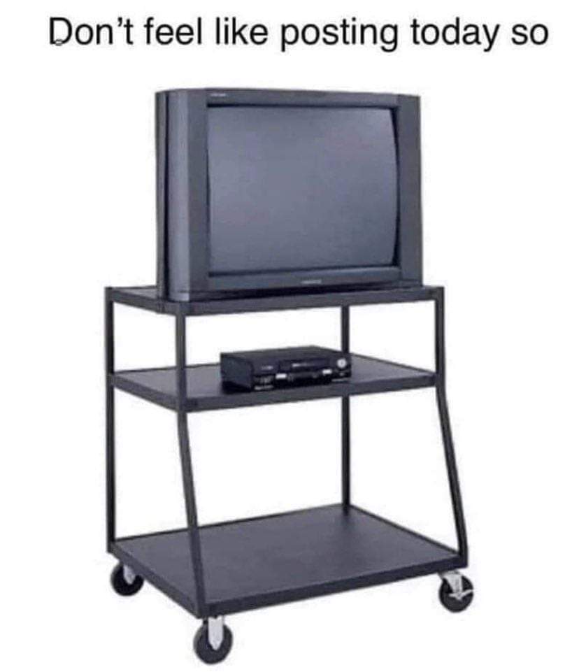 An image of a black, three-tiered TV cart with wheels, holding an old CRT television on the top shelf and a VCR on the…