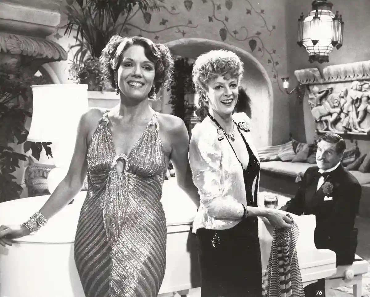 A black-and-white photo from a 1960s film shows two smiling women in glamorous evening wear standing beside a piano, while a…