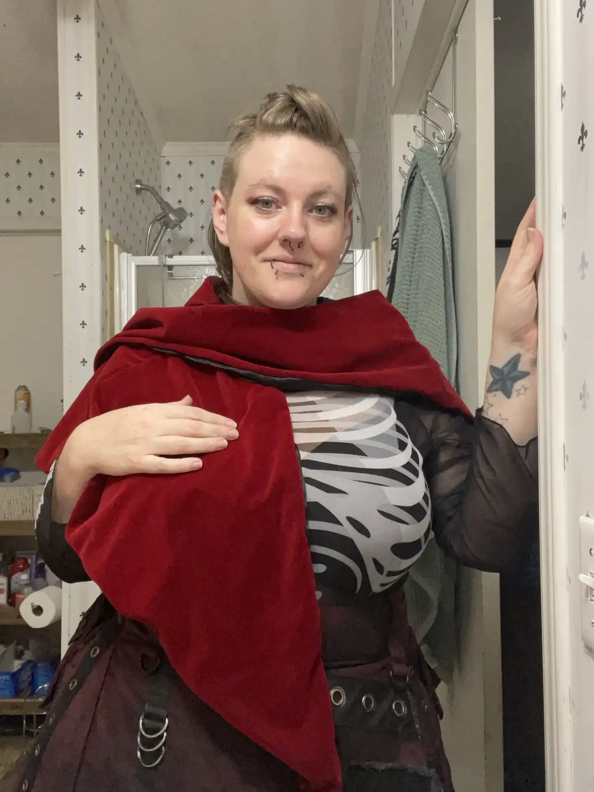 A person with multiple facial piercings and a star tattoo on their wrist stands in a bathroom doorway, wearing a red velvet…
