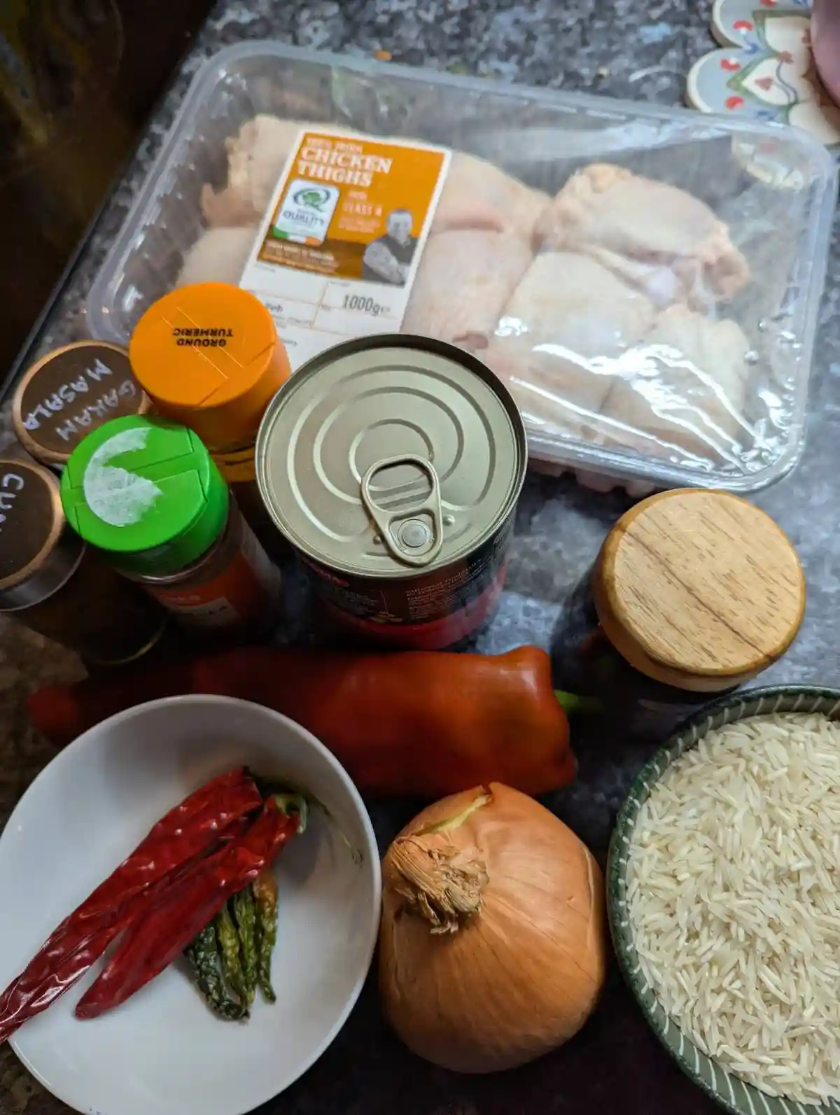 An overhead shot of ingredients for cooking, including a plastic tray of raw chicken thighs, a can of tomatoes, a bowl of…