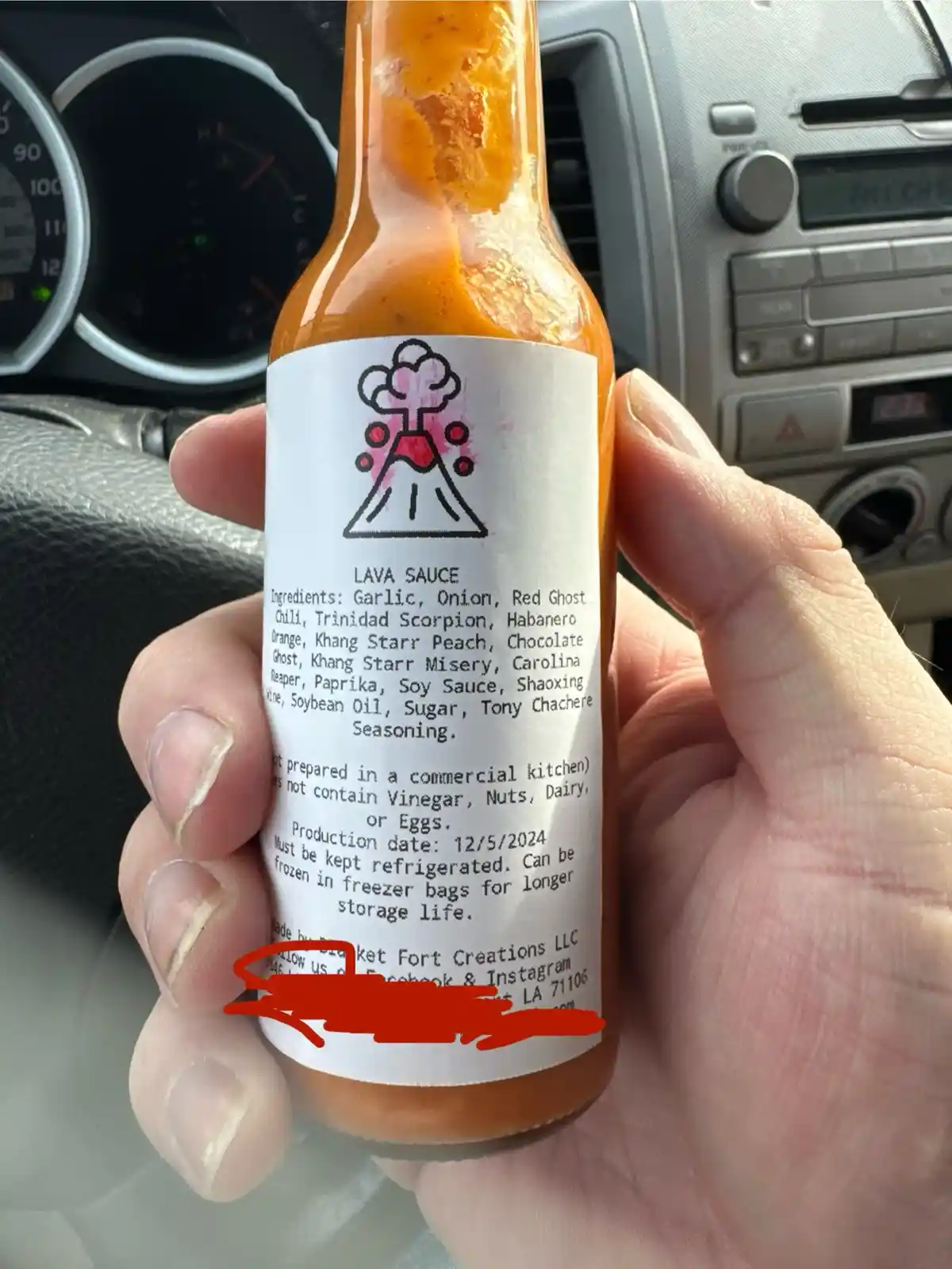 A hand holds a bottle of orange sauce inside a car, with the car's dashboard visible in the background. The label on the…