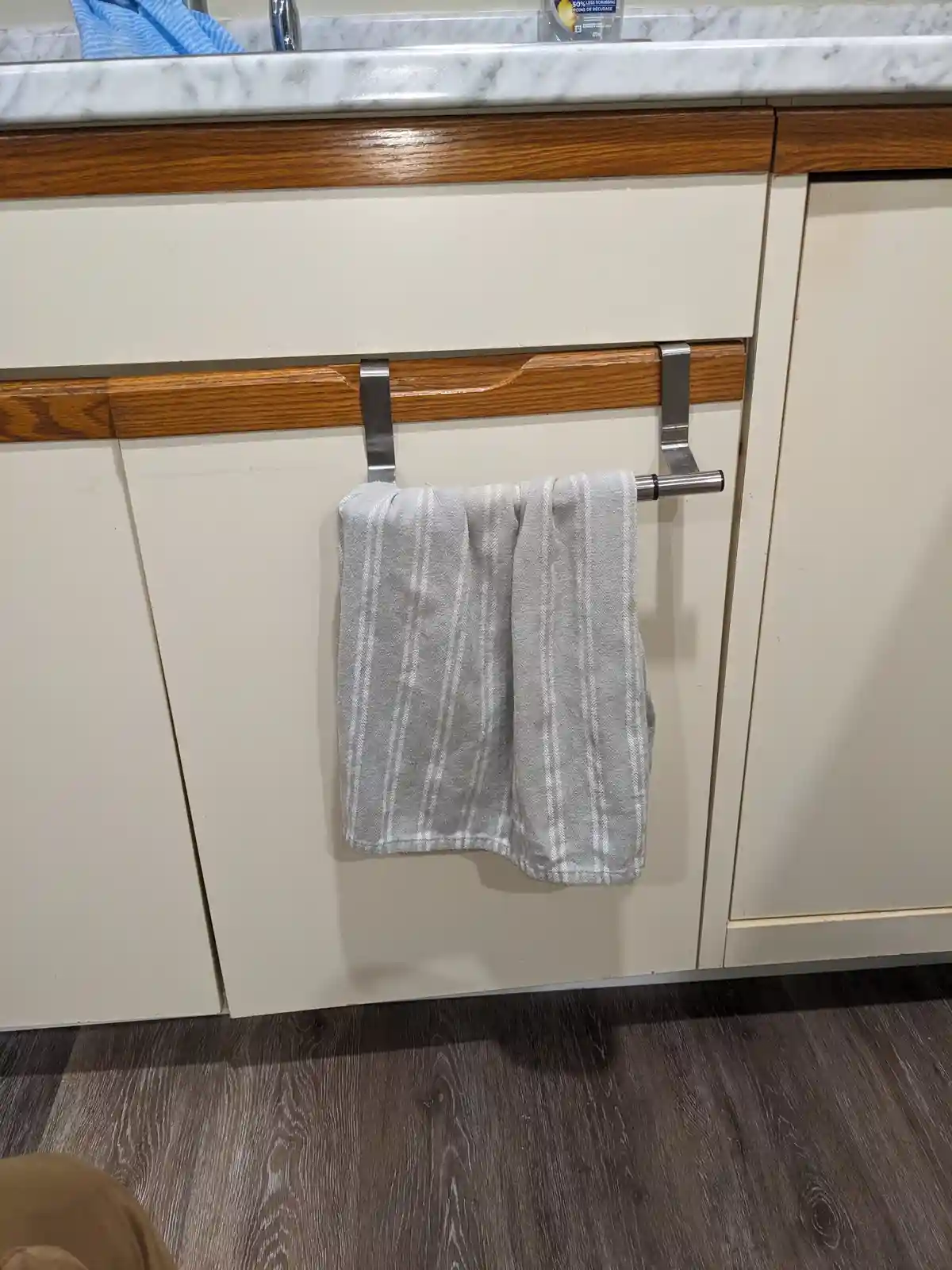 A gray and white striped towel hangs from a stainless steel over-the-door towel rack attached to a wooden cabinet handle…