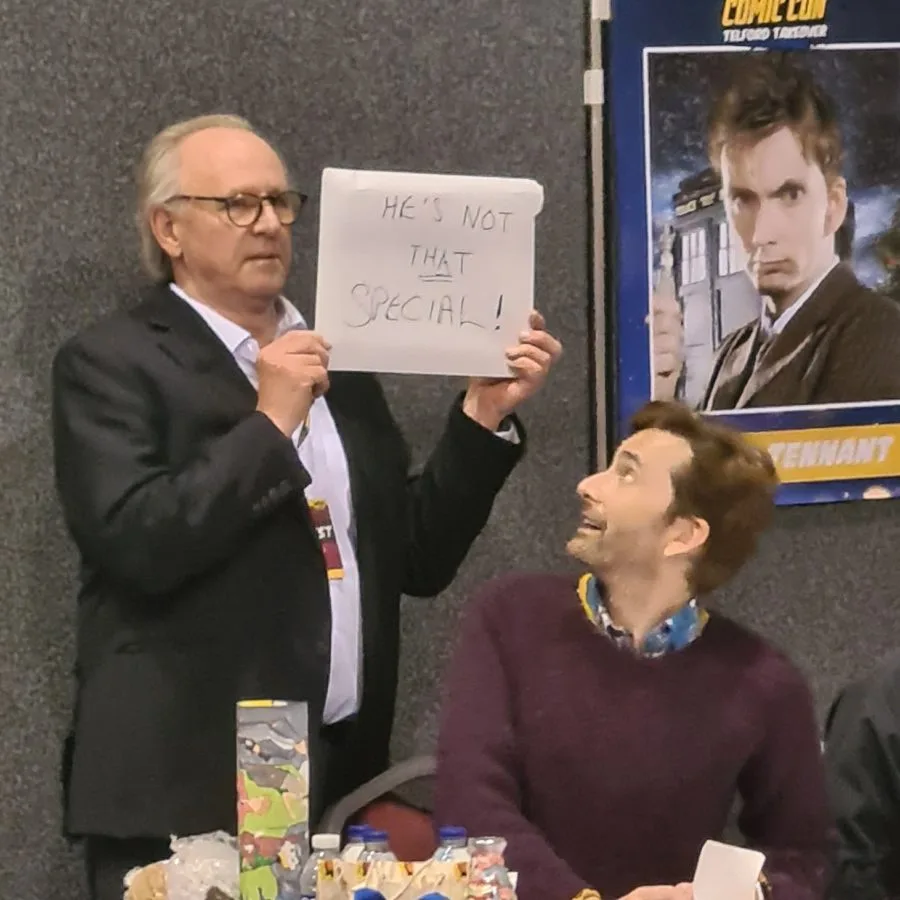 A man in a suit holds a handwritten sign reading 'HE'S NOT THAT SPECIAL!' while looking at another man seated at a table…