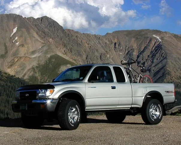 A silver Toyota Tacoma pickup truck is parked on a gravel road with a mountainous, rocky landscape and blue sky with clouds…
