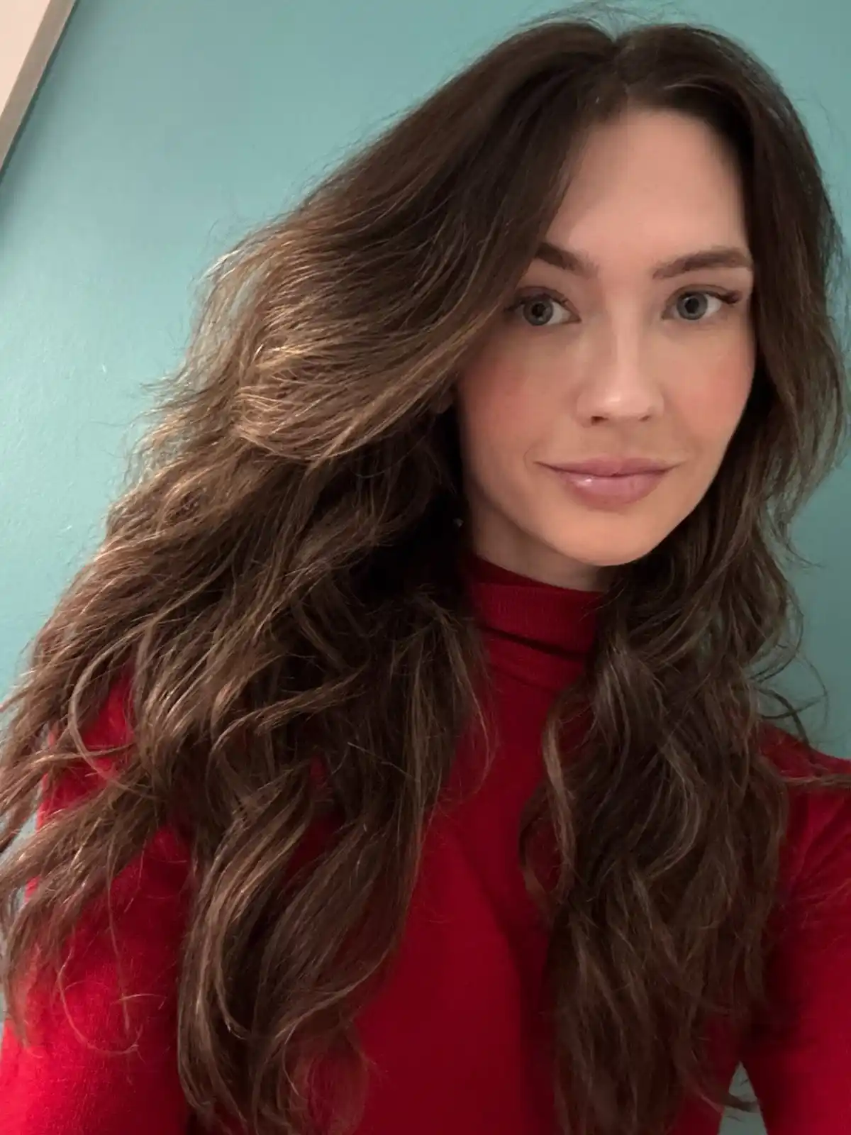A selfie of a woman with long, wavy brown hair and light eyes, wearing a red turtleneck against a teal wall. The overlaid…