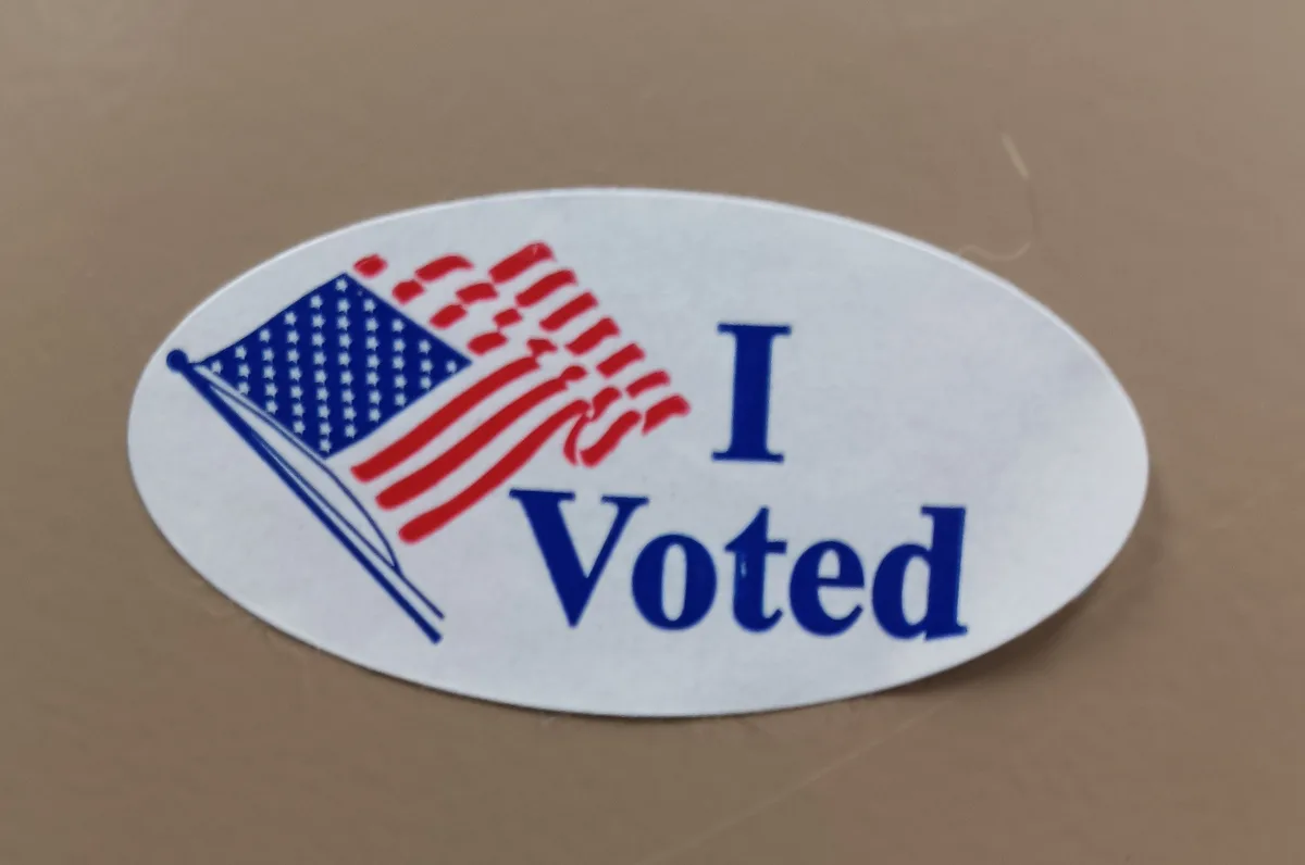 A close-up photo shows a white oval sticker with a stylized American flag on the left and the text 'I Voted' in blue…