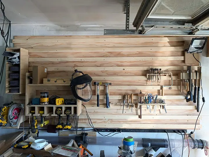 An overhead view of a well-organized garage workshop featuring a large wooden wall with neatly arranged tools including…