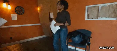 A woman in a dark shirt and jeans holds papers and walks toward a doorway in an orange-walled room with a dartboard on the…