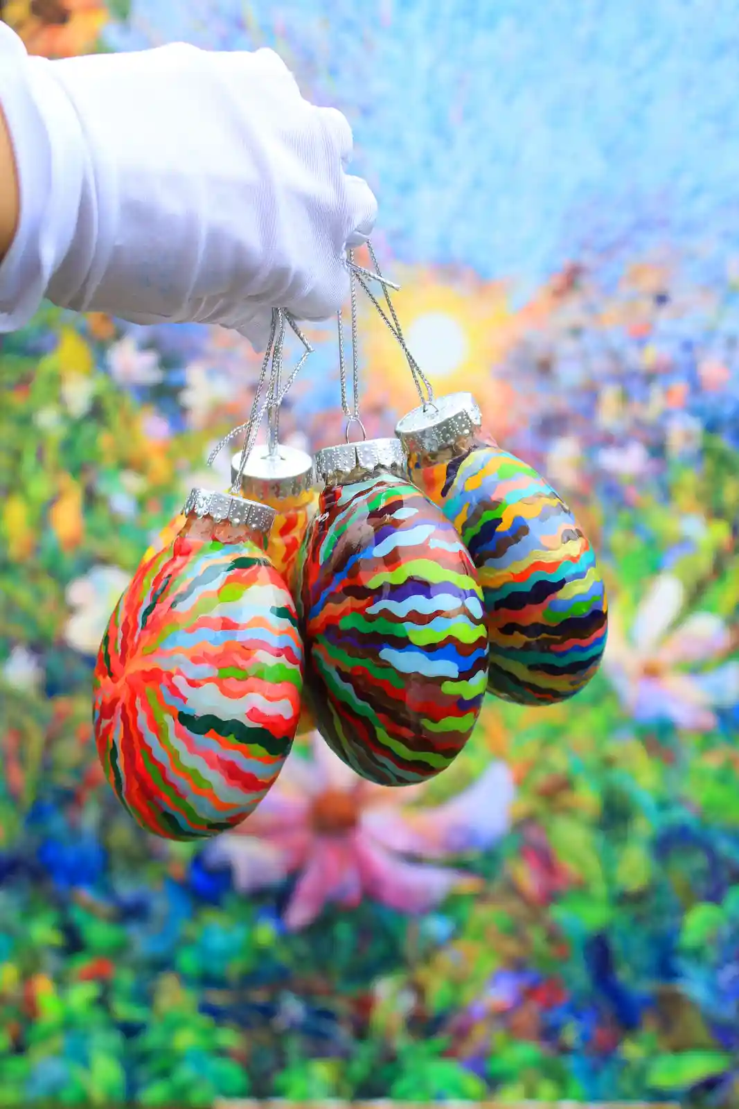 A white-gloved hand holds four colorful, wavy-striped Christmas ornaments against a blurred, vibrant floral background. The…