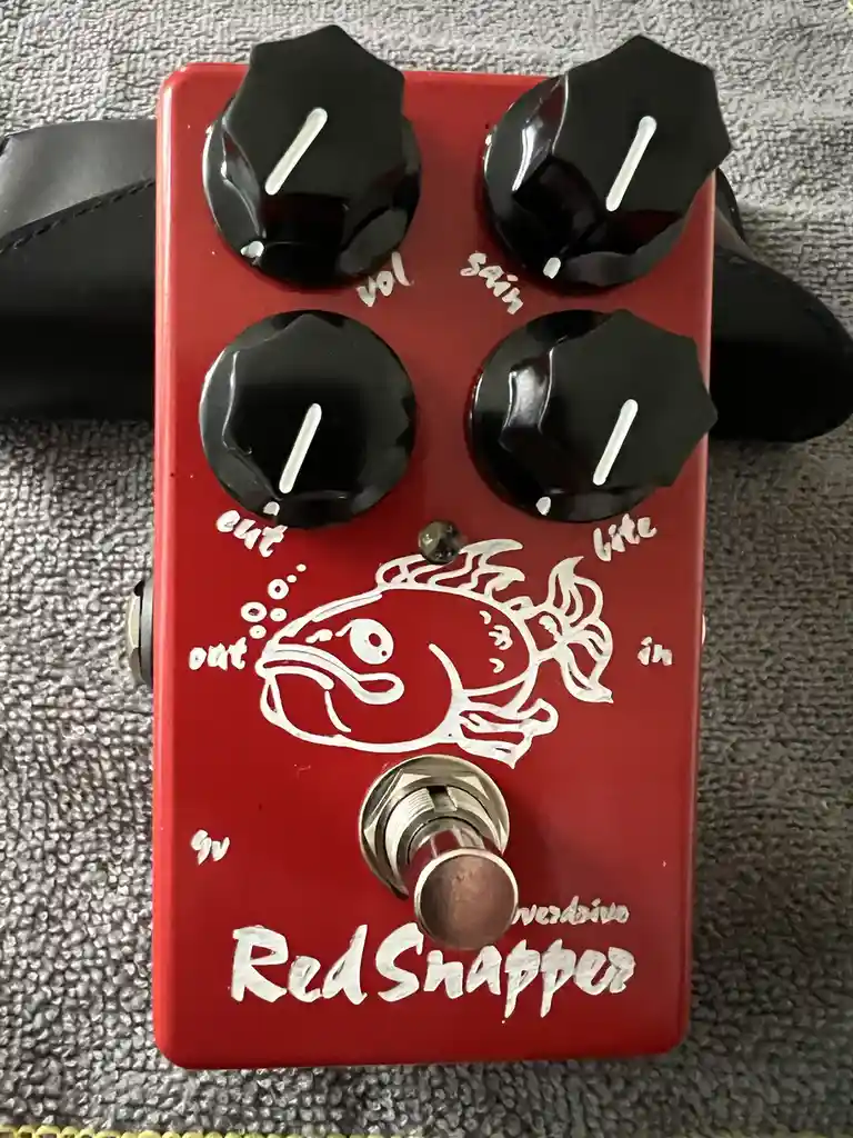 A red guitar effects pedal labeled 'RedSnapper' with a white fish graphic, four black knobs labeled 'vol', 'gain', 'out'…