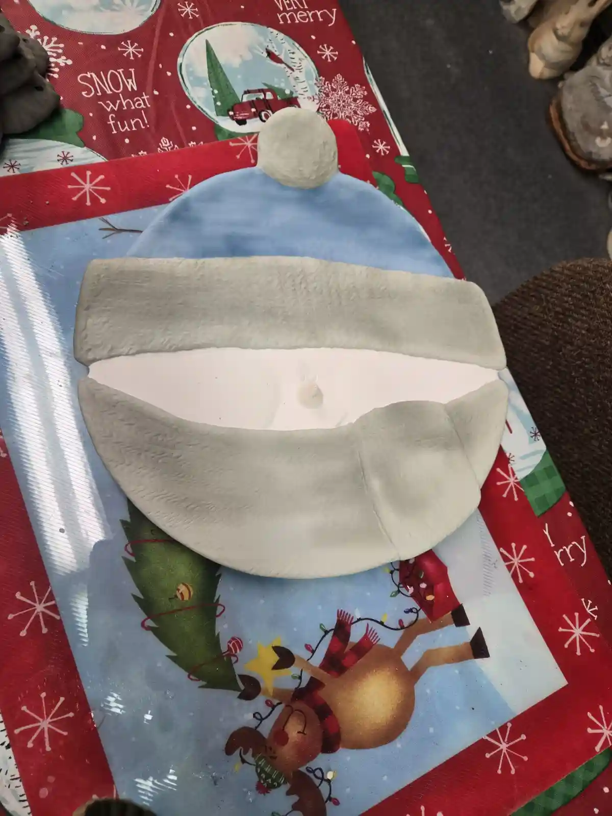 A handmade clay Santa hat, shaped like a bowl with a white interior and grey trim, sits atop a festive Christmas-themed…