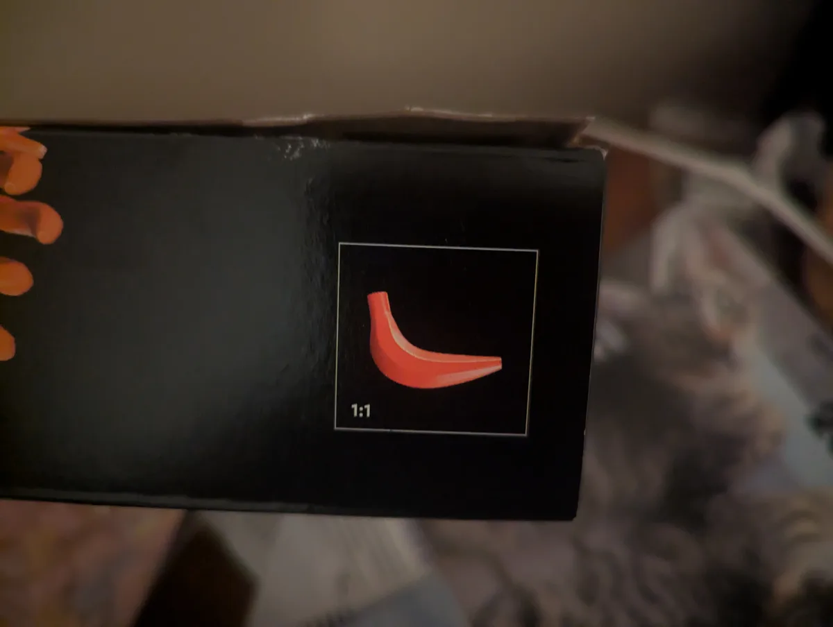 A close-up photo shows a black product box with a small square image on it, depicting a red, curved, crescent-shaped object…