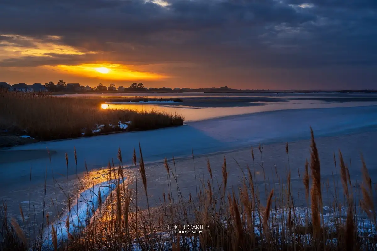 A serene winter sunset over a partially frozen body of water, with golden light reflecting off ice and reeds in the…