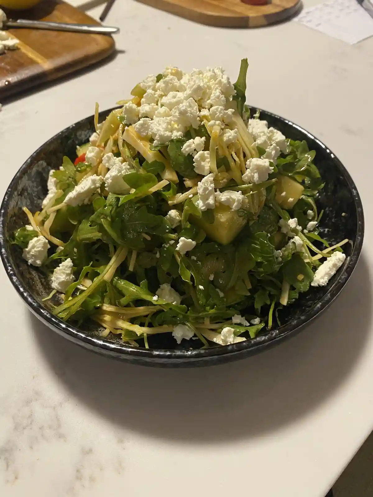A close-up photo shows a dark bowl filled with a salad of arugula, shredded vegetables, and crumbled white cheese, sitting…