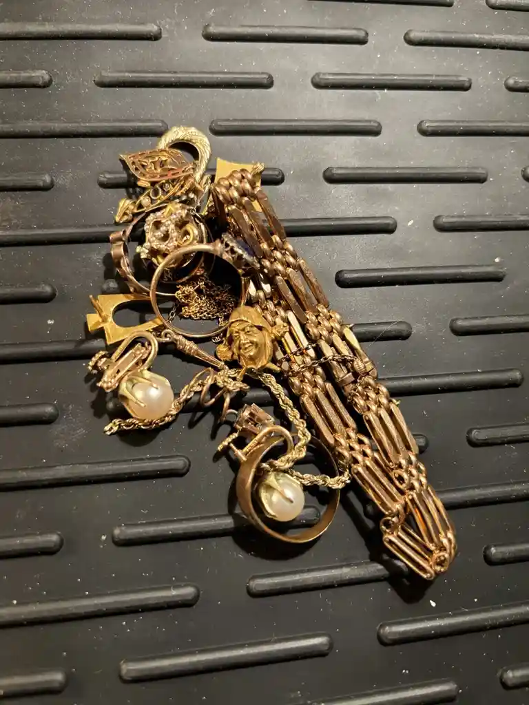 A pile of old, tarnished gold jewelry including chains, rings, a face charm, and pearl settings rests on a black rubber mat…
