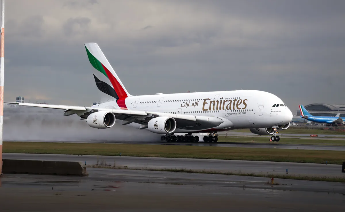 A large Emirates Airbus A380 airplane is shown on a wet runway, its landing gear touching down and creating a significant…