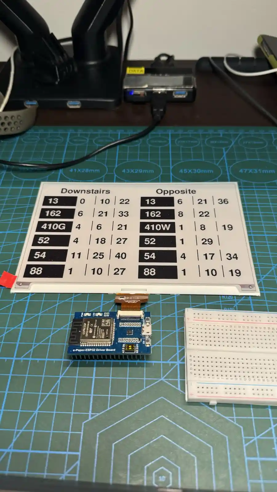A photo of an e-paper display showing two columns labeled 'Downstairs' and 'Opposite', each with rows of numbers and codes…