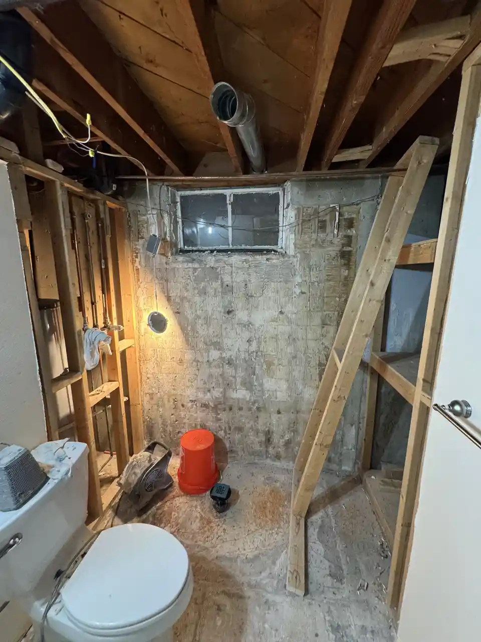 A bathroom under renovation shows exposed wooden beams, concrete walls, and a toilet with no surrounding fixtures. An orange…