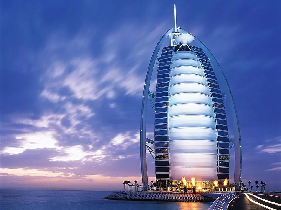 A stunning photograph of the Burj Al Arab hotel in Dubai at dusk, illuminated against a twilight sky with streaky clouds…