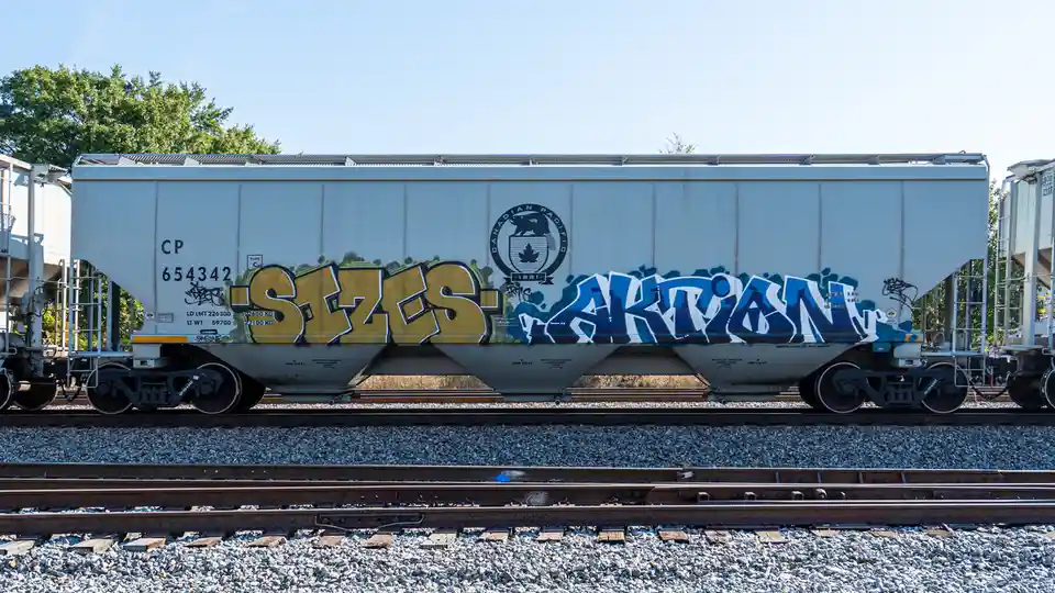 A white Canadian Pacific railcar sits on tracks under a clear sky, featuring large graffiti tags reading 'SIZES' in yellow…