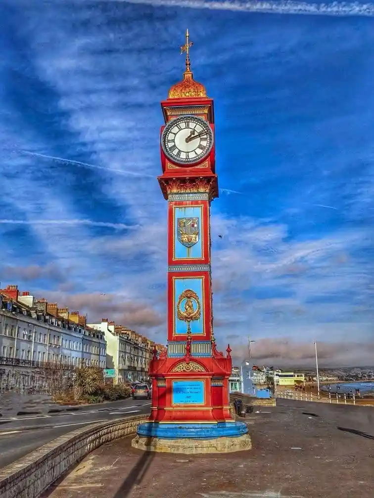 A tall, ornate red and blue clock tower stands prominently against a vivid blue sky with wispy clouds. The tower features a…