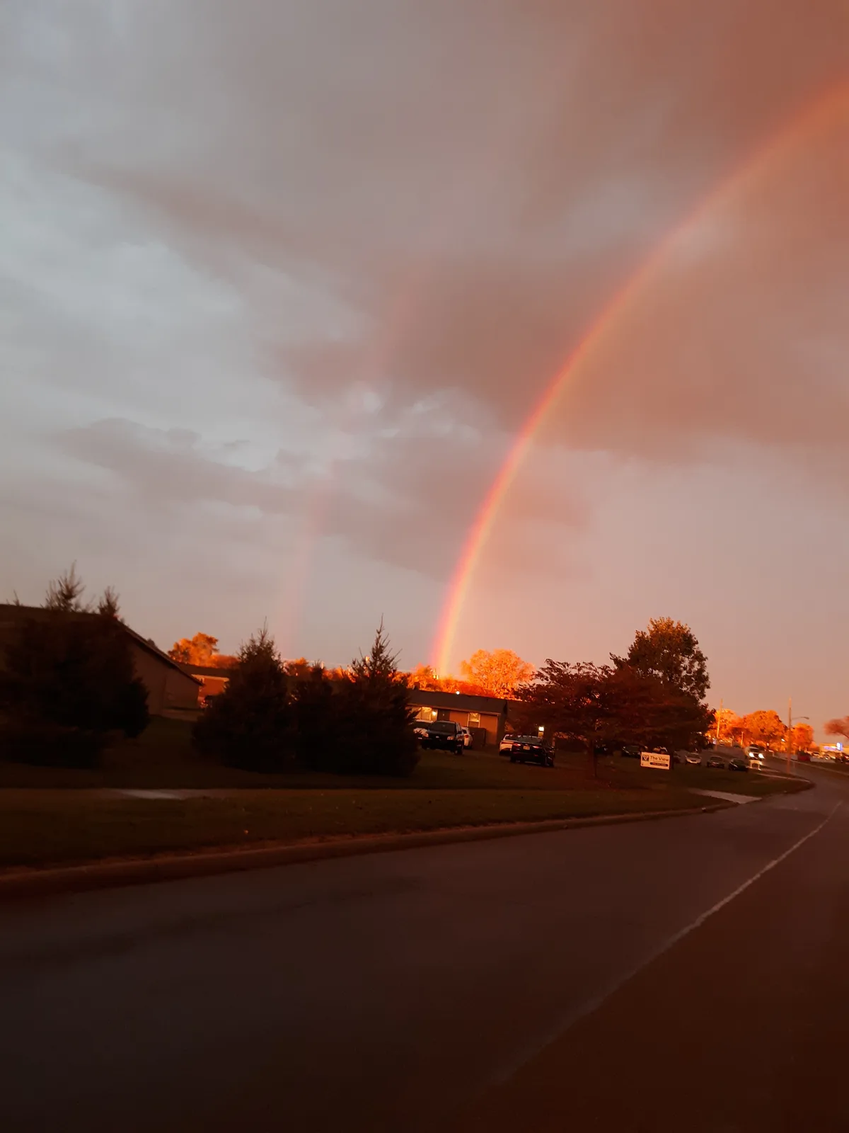 A photograph of a residential street at dusk or dawn, featuring a prominent, vividly colored rainbow arcing across a cloudy…