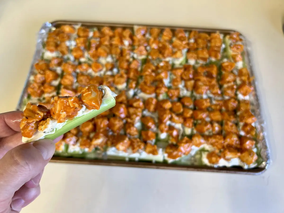 A hand holds a celery stick loaded with creamy white dip and orange buffalo chicken pieces, with a large baking sheet behind…