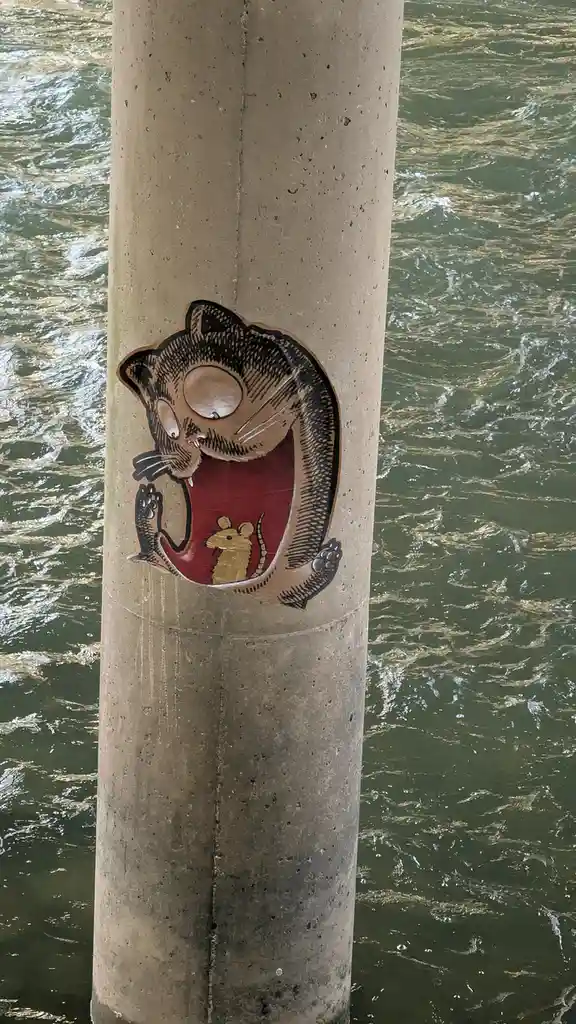 A cartoon-style sticker of a cat with its mouth wide open, holding a small mouse inside, is affixed to a concrete pillar…