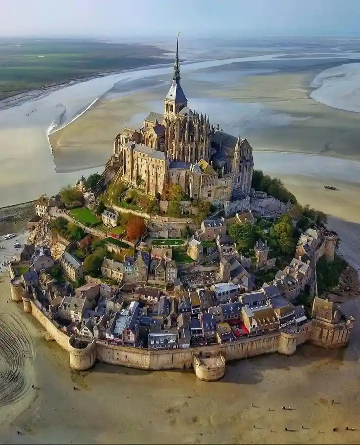 an aerial photograph shows the historic Mont Saint-Michel abbey and town perched atop a rocky island, surrounded by vast…