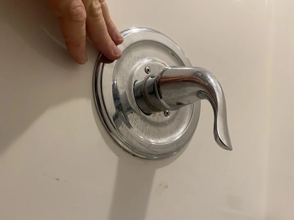 A close-up photo shows a person's hand gripping a chrome bathtub faucet handle, which is visibly dirty and worn. The faucet…