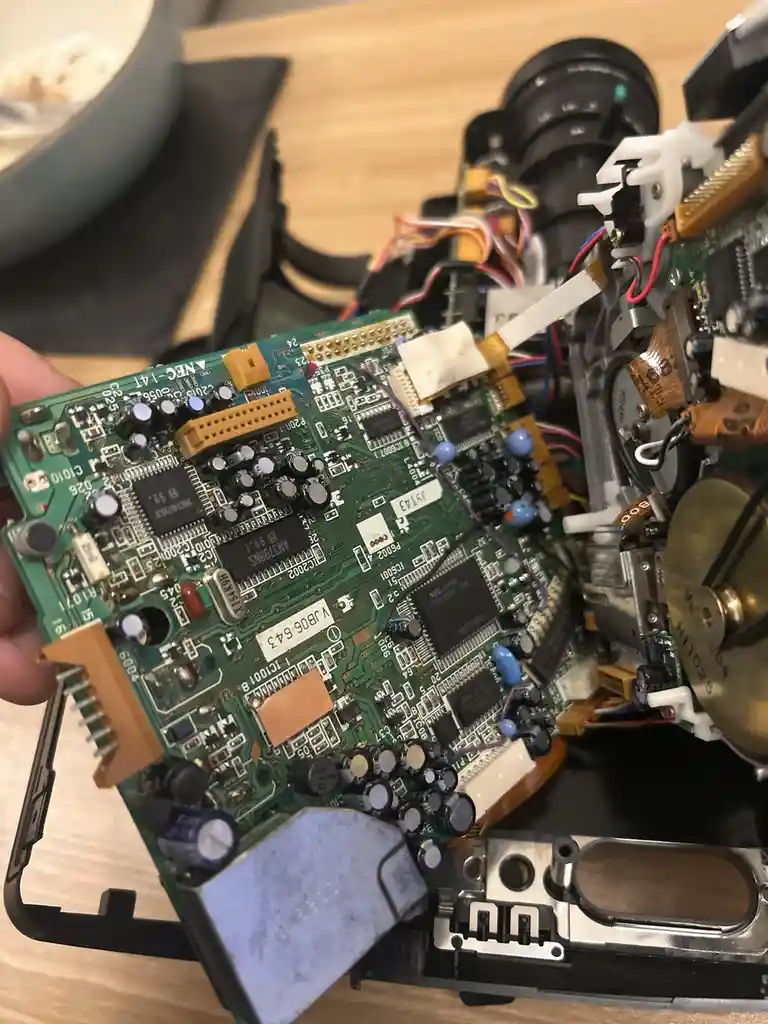 A close-up photo shows a hand holding a green circuit board from a disassembled camera, with its internal components like…