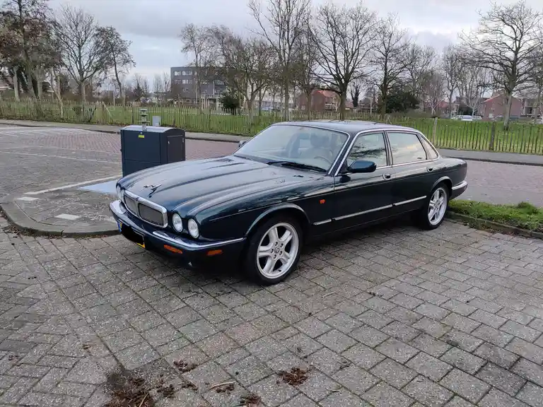 A dark green Jaguar XJ sedan is parked on a paved brick surface next to a sidewalk and a grassy area with bare trees. The…