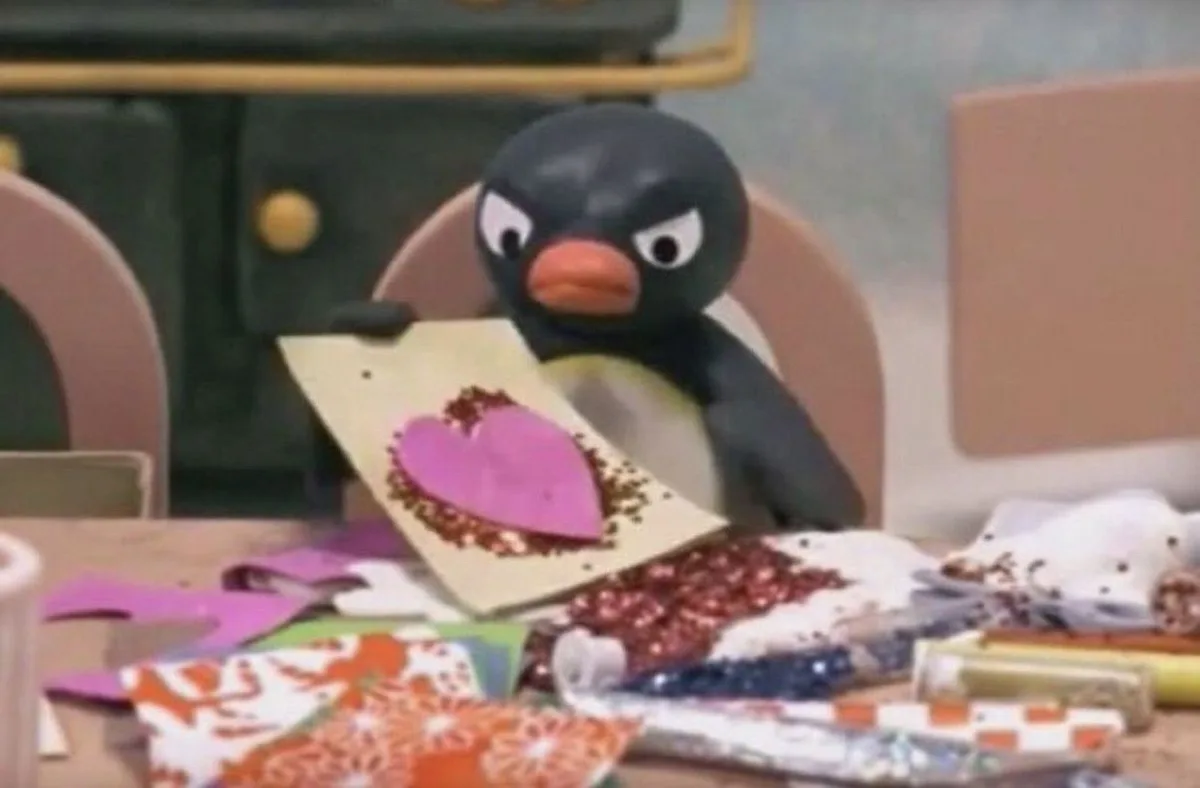 A penguin with wide, angry eyes sits at a cluttered craft table holding up a handmade card featuring a pink heart surrounded…