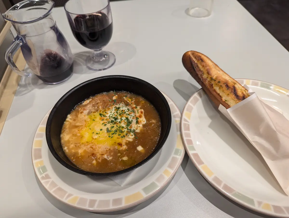 A bowl of soup with melted cheese and herbs sits on a patterned plate, next to a plate with a garlic breadstick and a glass…