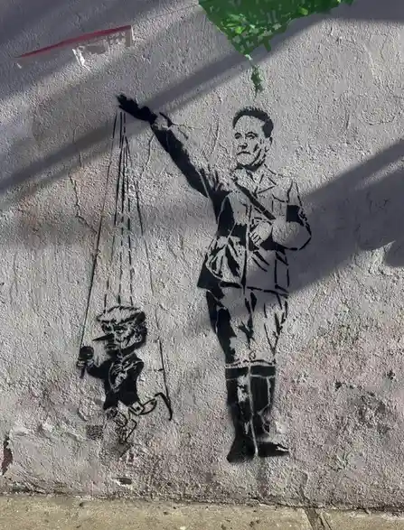 A black-and-white street art stencil on a textured wall depicts a man in a military-style uniform holding strings that…