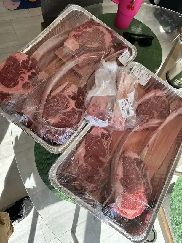 Two aluminum trays filled with large, raw, bone-in beef cuts, wrapped in plastic, sit on a glass table. A pink cup and…