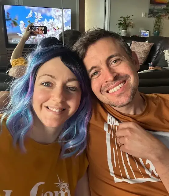 A smiling man and woman pose closely for a selfie in a living room. The woman has vibrant blue and purple dyed hair and…