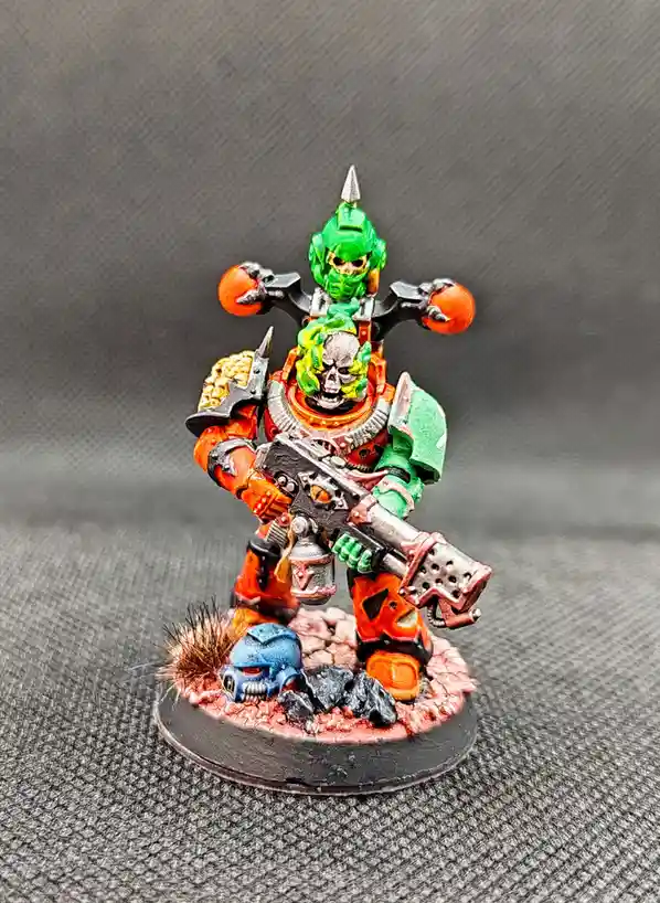 A painted miniature of a green-skinned, skull-faced warrior in orange and green armor, holding a large gun and two orange…