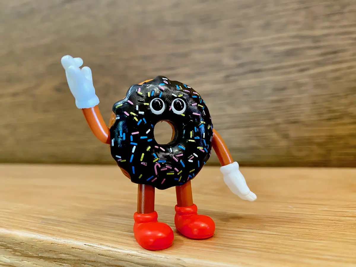 A small toy figurine shaped like a chocolate-frosted donut with colorful sprinkles, featuring large cartoon eyes, orange…