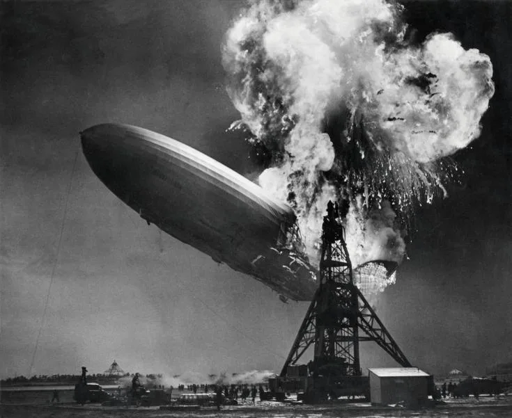 a black and white historical photograph shows the zeppelin airship LZ 129 Hindenburg engulfed in flames as it crashes onto a…