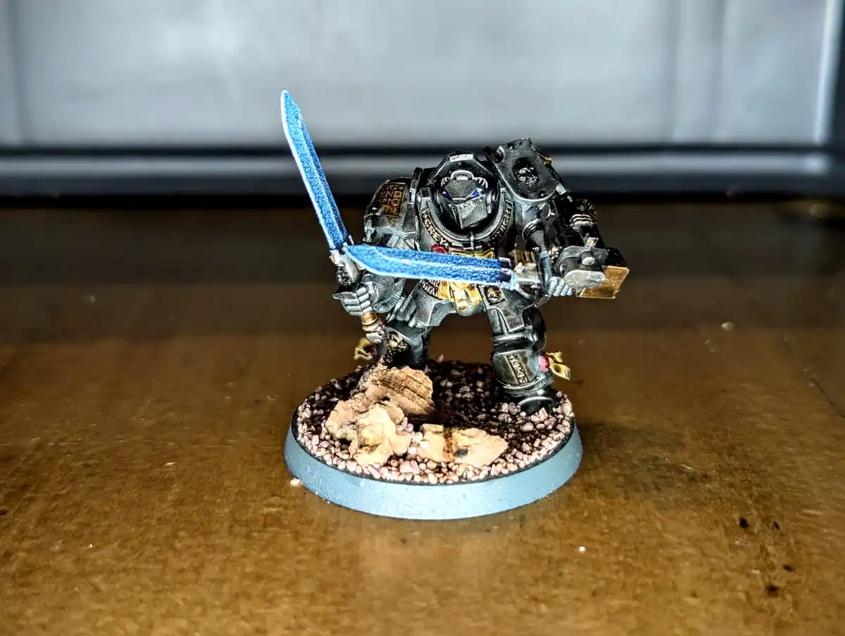 A painted miniature of a heavily armored, futuristic warrior with a glowing blue sword stands on a rocky base. The overlaid…