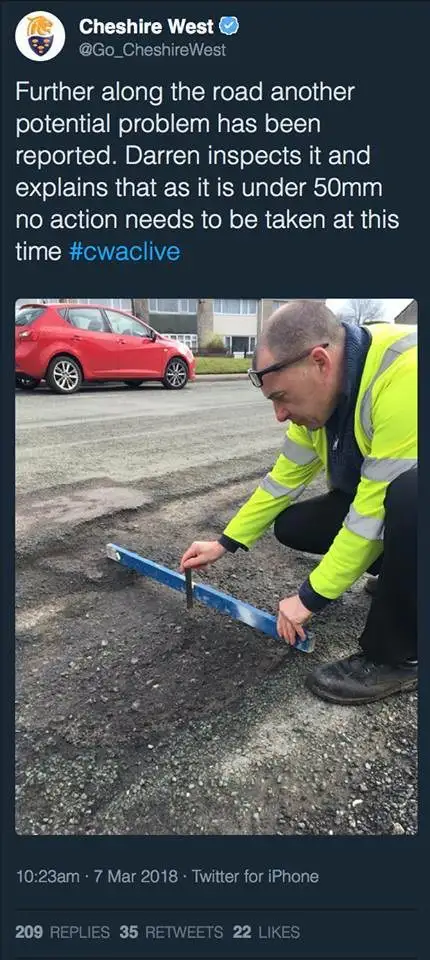 A man in a high-visibility jacket kneels on a road, using a blue ruler to measure a pothole. A red car is parked nearby…