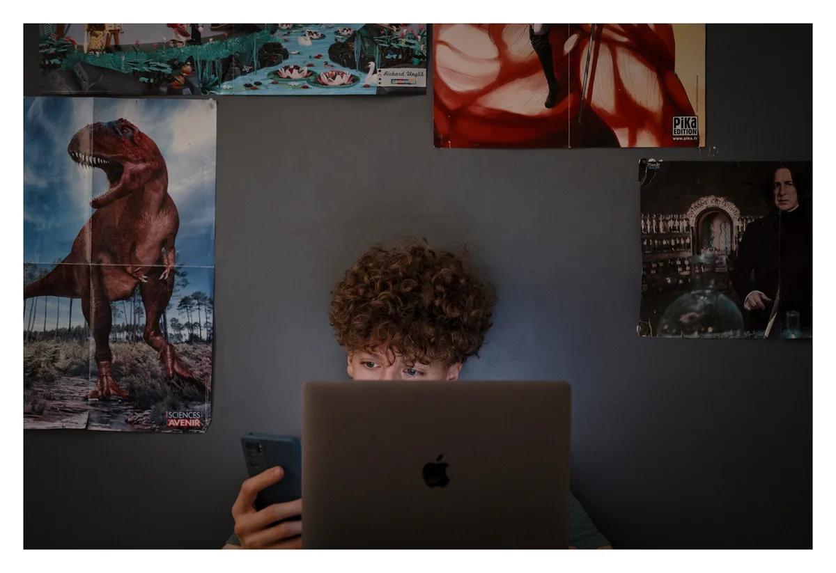 A person with curly hair is sitting at a desk, partially obscured by a laptop, looking intently at the screen while holding…