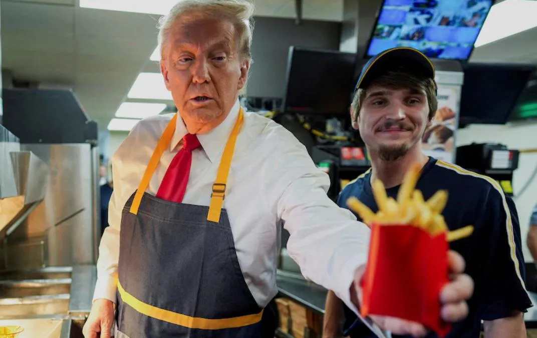 A man in a white shirt, red tie, and dark apron with yellow trim holds out a red paper container of french fries toward the…