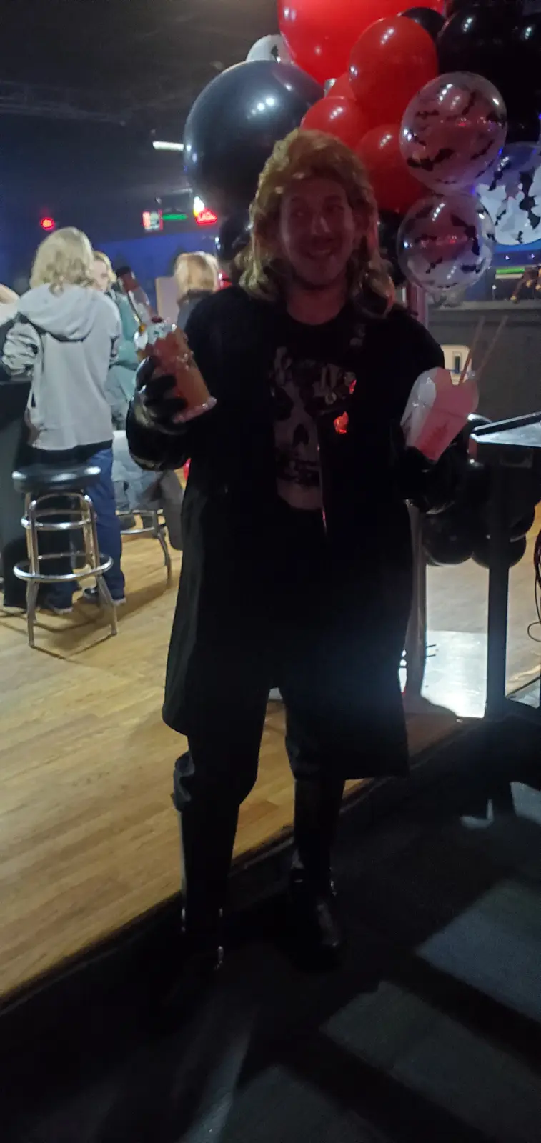 A person with long blonde hair, wearing a black outfit and black gloves, smiles while holding two drinks at a party with…