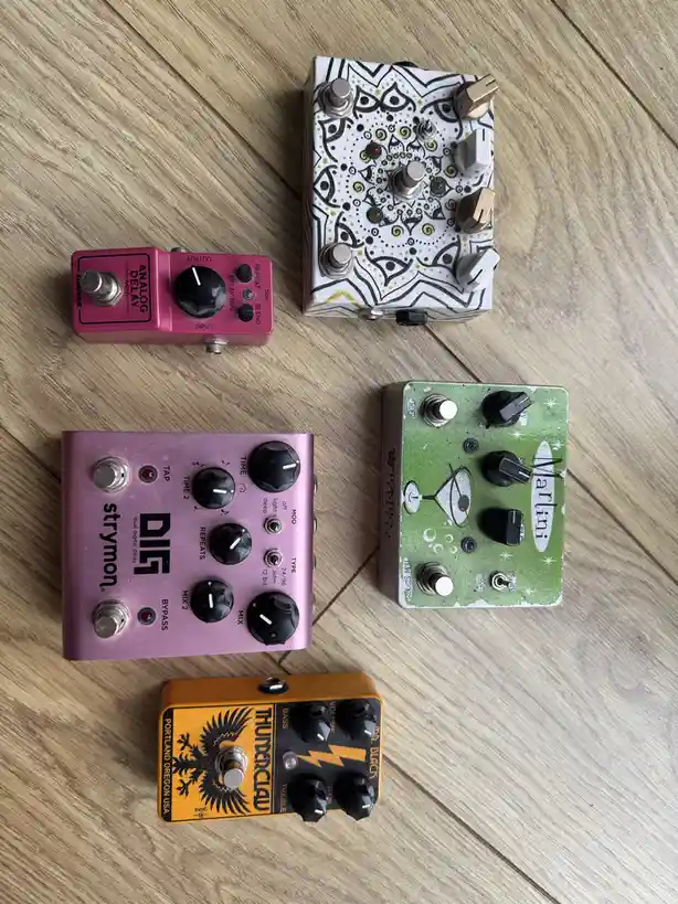 Five colorful guitar effect pedals are arranged on a wooden floor. A pink 'Analog Delay Mini' pedal, a white and black…