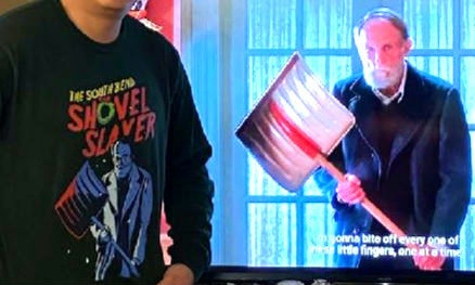 A man in a black hoodie with a graphic of a man holding a shovel and the text 'THE SOUTH BEND SHOVEL SLAYER' stands beside a…