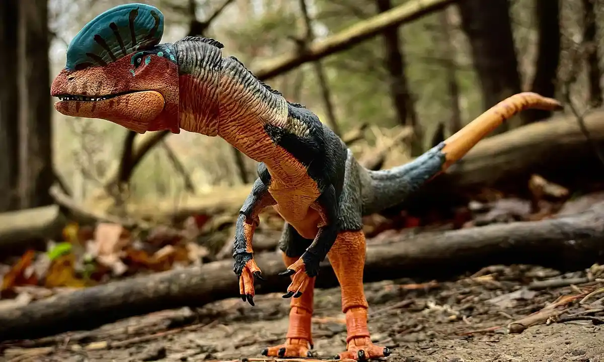 A detailed plastic toy dinosaur with a blue crest, orange and grey body, and black claws stands in a forest setting. The…
