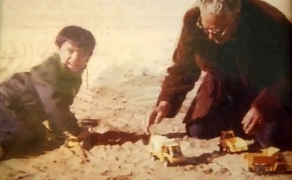 An old, grainy photo shows a young child and an older man, likely their father, kneeling on sandy ground playing with yellow…