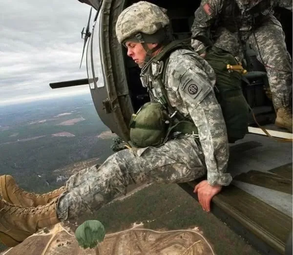 A soldier in full camouflage gear is seated at the open side of a helicopter, looking down at the ground far below. His body…
